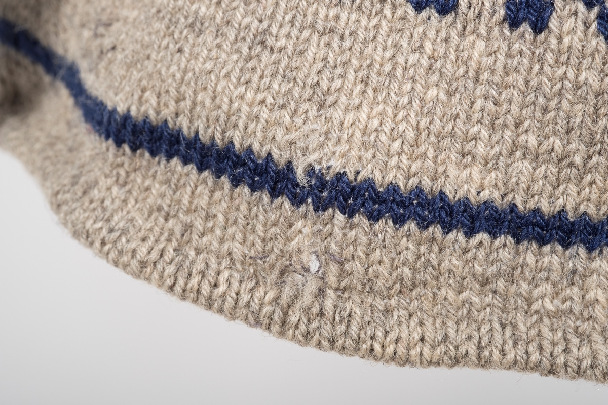 Blå og grå strikket herregenser. 
To knapper ved halsåpningen for å kunne kneppe den igjen. 
Ermekantene er brettet innover og sydd fast. 
To sømkanter på baksiden av genseren.