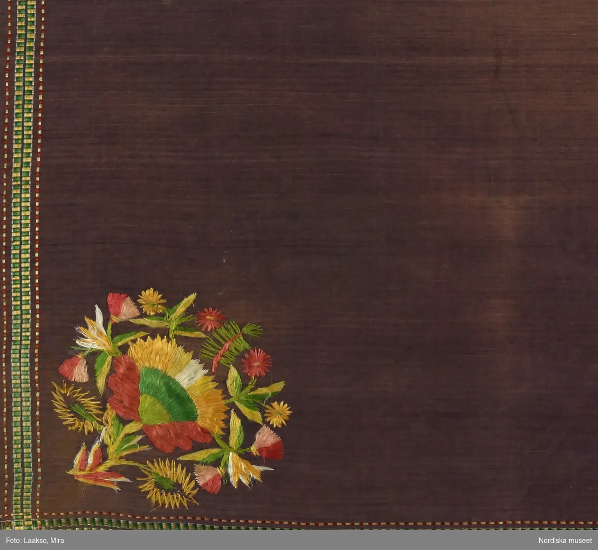 Kvadratiskt halskläde använt som bokkläde, av lila bomullslärft, enfärgat med en smal kantbård i rips med silke i gult, grönt och rött. . I ena hörnet  blommotiv broderat med otvinnat silke i rött, gult, grönt i plattsöm. Fint sydda smala fållar.
Anm. Ojämnt blekt.
Tillhör en grupp av likartade som även vävdes i siden från tidigt 1800-tal. Vanliga i södra Sverige och i Danmark. Finns även i t.ex. Härjedalen se inv.nr 103578 och från Medelpad  198.494.
/Berit Eldvik 2012-01-10