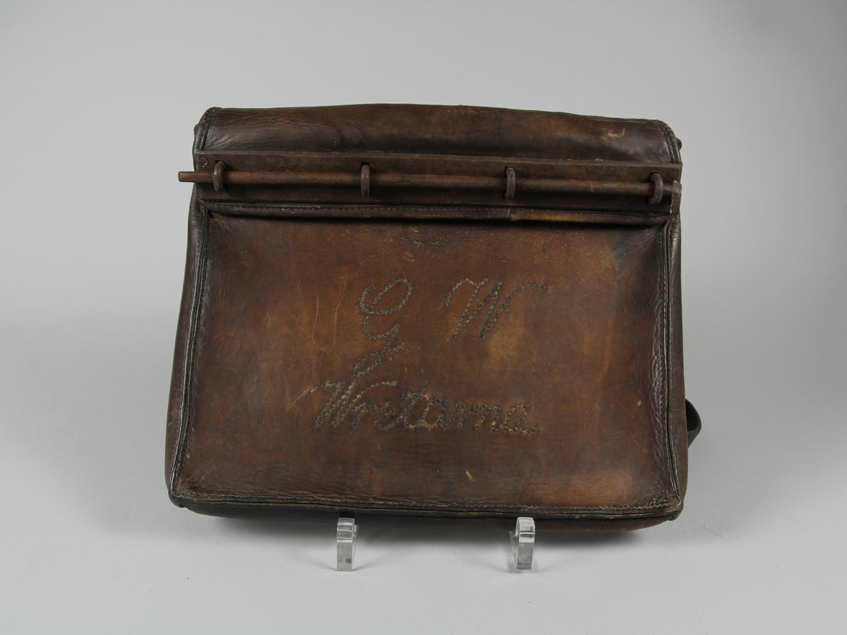 Postväska, rektangulär, av brunt skinn med reglerbar bärrem. Smal klaff/fällock som låses med fyra metallöglor och lång rund metallten.