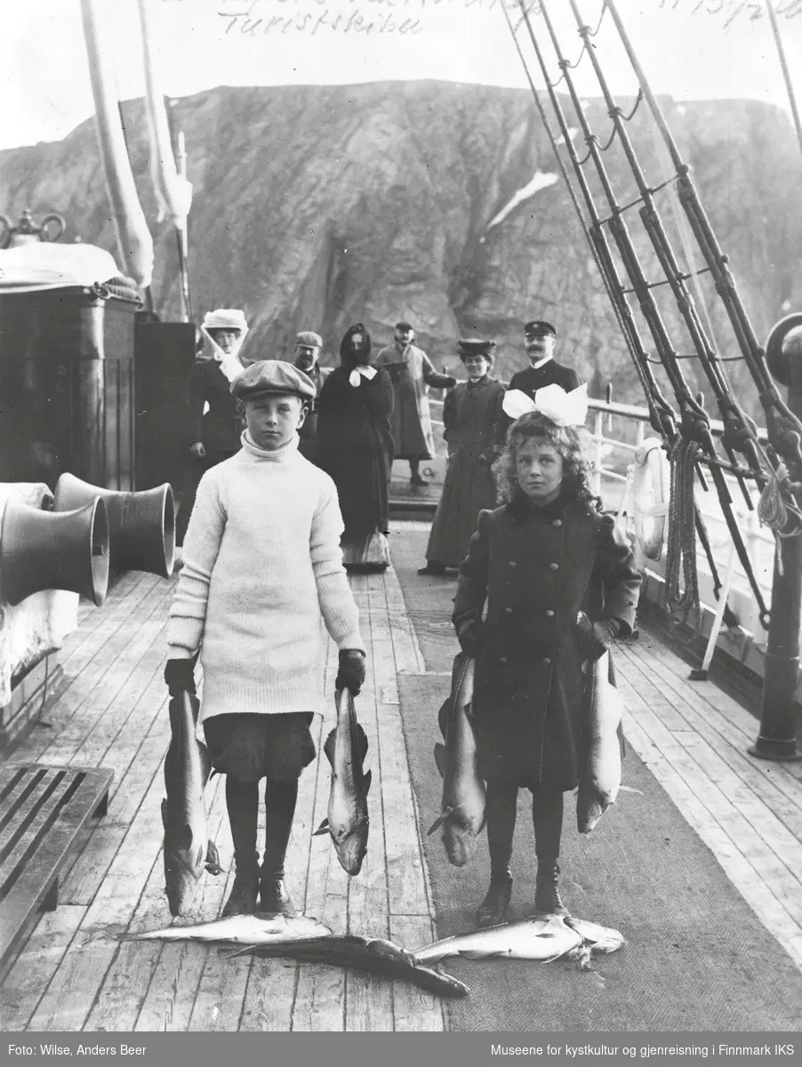 Passagerer ombord D/S "Bergensfjord" utenfor Nordkapp. I forgrunnen to barn med fisk. Juli 1926. (Dato etter "galleri NOR", men etter klesdrakt antas at bilde ble tatt mye tidligere.) - (Originalnegativet eies av Norsk Folkemuseum, W 28839*B)