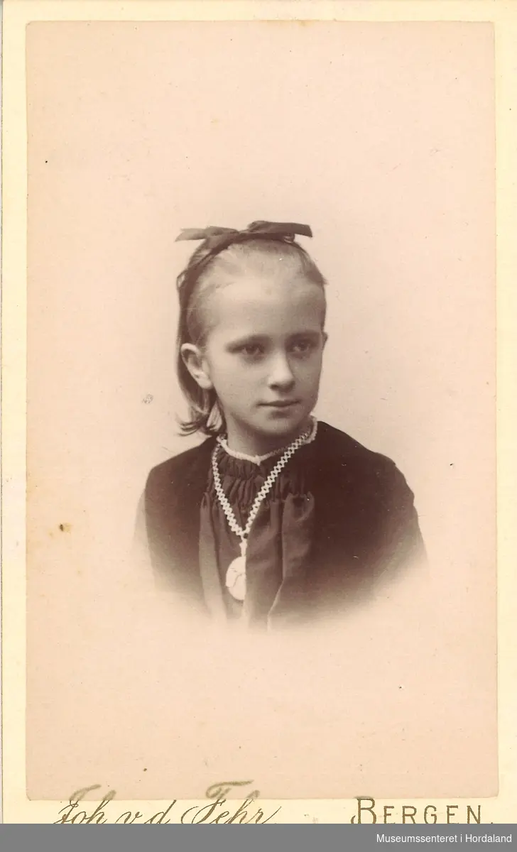 portrettfotografi av jente med mørk bluse/kjole med rynka brystparti. Håret samla med sløyfe oppe på hove, medaljong i kjede om halsen