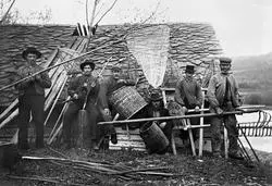 Fiskere, fiskeutstyr, fiskebu, lagesildsfiske, lågåsildfiske, fiske etter lågåsild. Bildet viser fiskere med forskjellig utstyr utenfor Knuvelbua i 1904.  Bildet er fra Knuvelen (Kringelen) i Fåberg (i nyere tid Lillehammer kommune). Knuvelbu. Knøvelbu. Knøvelbua. Knuvelbua. Gudbrandsdalslågen.

Tore Johan Brevik opplyste i en e-post til museet 22. oktober 2015 at mannen helt til høyre på fotografiet er hans oldefar på morssida, Hans Olson Vold, og at mannen bak ham (nest ytterst til høyre) er bestefaren, Syver Kristiansen Solbakken.

Knuvelbua ble flyttet til friluftsmuseet på Maihaugen i 1921.  Museumsgründeren Anders Sandvigs omtale av bygningen er gjengitt under fanen «Opplysninger».