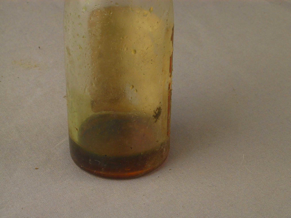 Glasflaske med jamn overgang mellom korpus og hals. Etikett. Inneheld liten skvett med olje,  "kork" av papir.