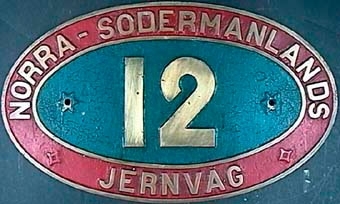Lokskylt av mässing från Norra Södermanlands Jernväg. Märkt 12 och målad i rött och blått.