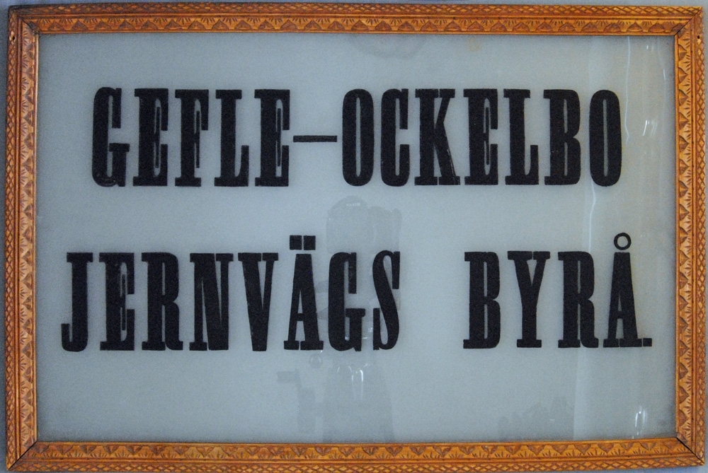 Skylt som suttit vid "Gefle-Ockelbo Jernvägs Byrå"
Ram i snidat trä, tryckta svarta bokstäver på etsat glas.