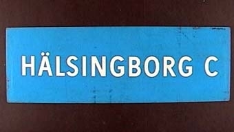 "Rektangulär dubbelsidig plastskylt med vit text på blå botten: 
HÄLSINGBORG C".

På andra sidan:
"HÄSSLEHOLM".