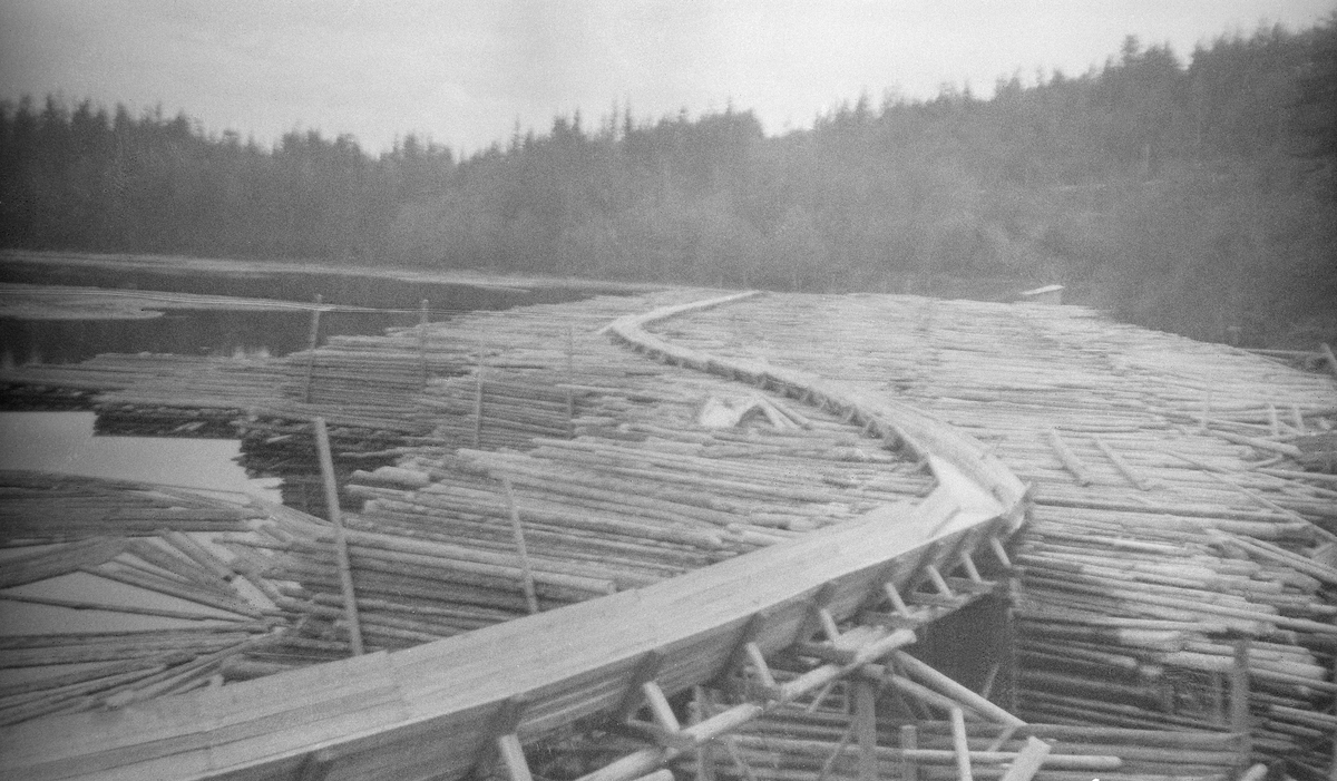 Tømmerrenne og tømmeropplag ved papirfabrikken Follum.  Fotografiet er tatt på et sted der elva Begna løper inn mot en stillestående sjø i Begnavassdraget, antakelig like ovenfor den nevnte papirfabrikken.  På strendene i forgrunnen lå det ei kjempediger lunnevelte.  Tømmeret var stablet tett oppunder den nevnte tømmerrenna, som slynget seg over velta.  Renna var lagd av planker, og hadde tømmerløp med trapesformet tverrsnitt.  I forgrunnen kan det synes som om det var en forholdesvis åpen passasje i tømmervelta, antakelig for å gi rom for elvas hovedløp på dette stedet.

I 2017 fikk Norsk Skogmuseum en e-post fra Thor Henriksen, som hadde sett dette fotografiet på nettstedet DigitaltMuseum.  Han hadde følgende kommentar:

«Dette bilde er nok tatt på nedsiden av Ådal tresliperi som lå på vestsiden av Hensfossen i Ådalselva.
Tresliperiet brant under andre verdenskrig og i dag ligger det en kraftstasjon der.
Tømmeret i vika skulle inn til tresliperiet. Vika ble forøvrig fylt igjen med bark fra Follum fabrikker på 60 - 70 tallet.Til høyre kan man skimte Randsfjordbanen»