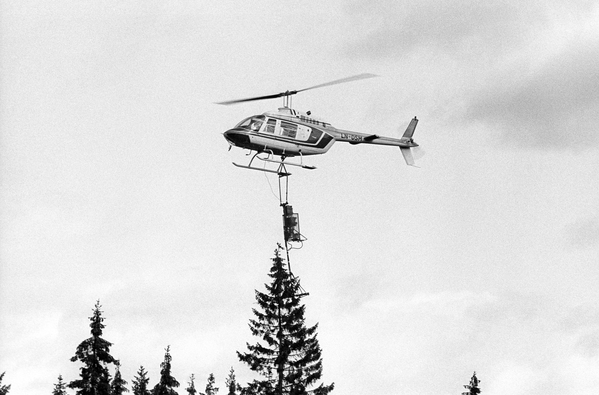 Helikopter med utstyr for «toppklipping», avbildet idet piloten var i ferd med å manøvrere apparatet han hadde hengende under seg mot en av de større grantoppene i skogen under seg.  Fotografiet er tatt i nærheten av Sigernessjøen i Kongsvinger, der det i 1983 ble foretatt konglehøsting fra et såkalt «elitebestand» ved hjelp av dette helikopteret.  Norsk institutt for skogforskning (NISK) og Skogfrøverket hadde latt to medarbeidere reise landet rundt for å registrere slike spesielt gode bestand gjennom 1970-åra med henblikk på at konglesankinga skulle orienteres mot områder med slik god skog inntil frøplantasjene med spesielt utvalgte kloner ble konglebærende.  Skogeierne skulle få godt betalt for de konglene som ble høstet fra deres «elitetrær».  Likevel var det langt fra alle som ønsket å hogge slike bestand når det ble frøår.  For å skape et alternativ til risikobetont og tidkrevende klatring på slike lokaliteter utviklet piloten Georg Strømme utstyr som skulle gjøre det mulig å «klippe» toppene av grantrærne i elitebestandene.  Det var nemlig i toppene størstedelen av konglene satt.  De hadde slik sett en verdi for Skogfrøverket, men hadde knapt noen verdi for skogeierne fordi de uansett ikke ville utvikle seg til salgbart virke når trærne skulle felles noen år seinere.  Helikopterutstyret ble prøvd første gang i Hedmark høsten 1983.  Det besto av en innretning som hang under helikopteret, som ble manøvrert slik at den omsluttet tretoppene som skulle kappes.  Når utstyret var i riktig posisjon kunne en utløse et hydraulisk slag, som knekte toppen slik at den falt i bakken. 

Dette var de første forsøkene med toppklipping med sikte på konglesanking i Norge.  Nye forsøk ble gjort i 1987-88, da med et kraftigere aggregat som ingeniør Torstein Lisland fra driftsteknisk avdeling ved Norsk institutt for skogforskning (NISK) hadde utviklet.  Firmaet Helilift A/S og piloten Georg Strømme var fortsatt Skogfrøverkets samarbeidspartnere i denne virksomheten.