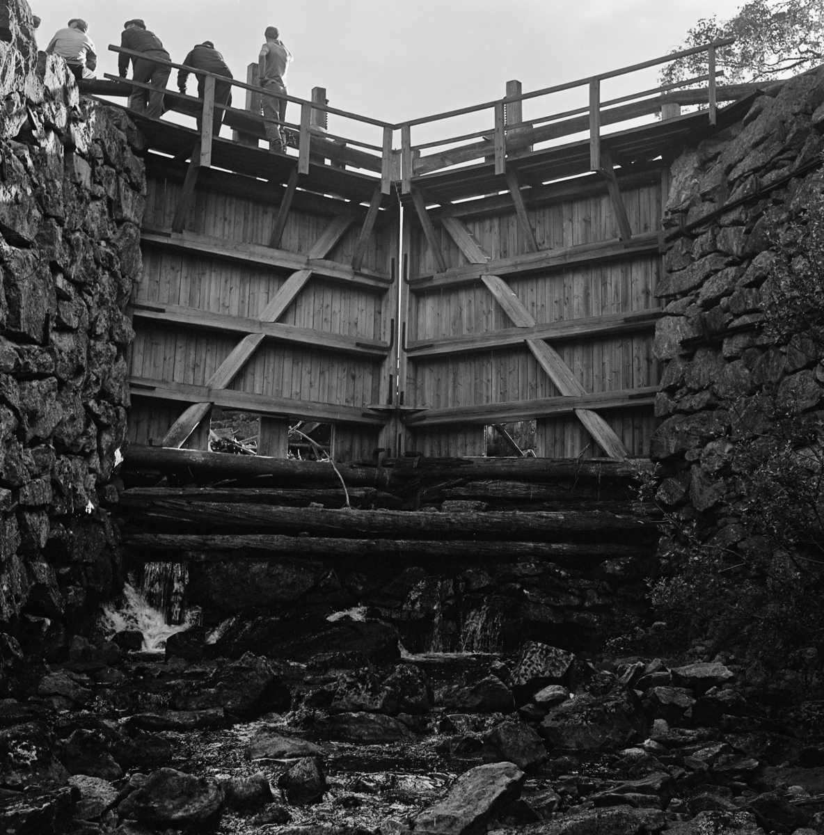 Sluseport i Grasmokanalen eller Sootkanalen i Eidskog, fotografert under ei befaring i 1978.  Fotografiet er tatt fri sjølve elveløpet, i motstrøms retning, altså opp mot porten.  Vassdraget ble kanalisert i slutten av 1840-åra, ble brukt til tømmertransport fram til begynnelsen av 1930-åra.  Det er vel grunn til å tro at den sluseporten som vises her er en seinere rekonstruksjon, lagd for å kunne formidle kanalen som kulturminne.  Noen av sluseportene ble skiftet ut med kopier av de opprinnelige i 1954, og dette kan være en av disse.  Terskelstokkene (nederst i forgrunnen) er sannsynligvis eldre.  Sluseportene er montert mot kraftige murte vanger av naturstein som er samlet i nærområdet.  Sluseportene er såpass brede at de møter hverandre som en forholdsvis butt «plog» i motstrøms retning.  De har et rammeverk av 8" X 8" firskårne bjelker og var kledd med tettstilte planker på motstrøms side (mot ovenforliggende slusekammer).  Konstruksjonen er avstivet med diagonale skråbjelker og med vinkeljern. På toppen av portene er det montert gangbruer med rekkverk.  Da dette fotografiet ble tatt sto det fire mennesker på brua over venstre port.  Botnlukene i sluseportene sto åpne da fotografiet ble tatt. 

En kort orientering om Grasmokanalens eller Sootkanalens historie finnes under fanen «Andre opplysninger». 
