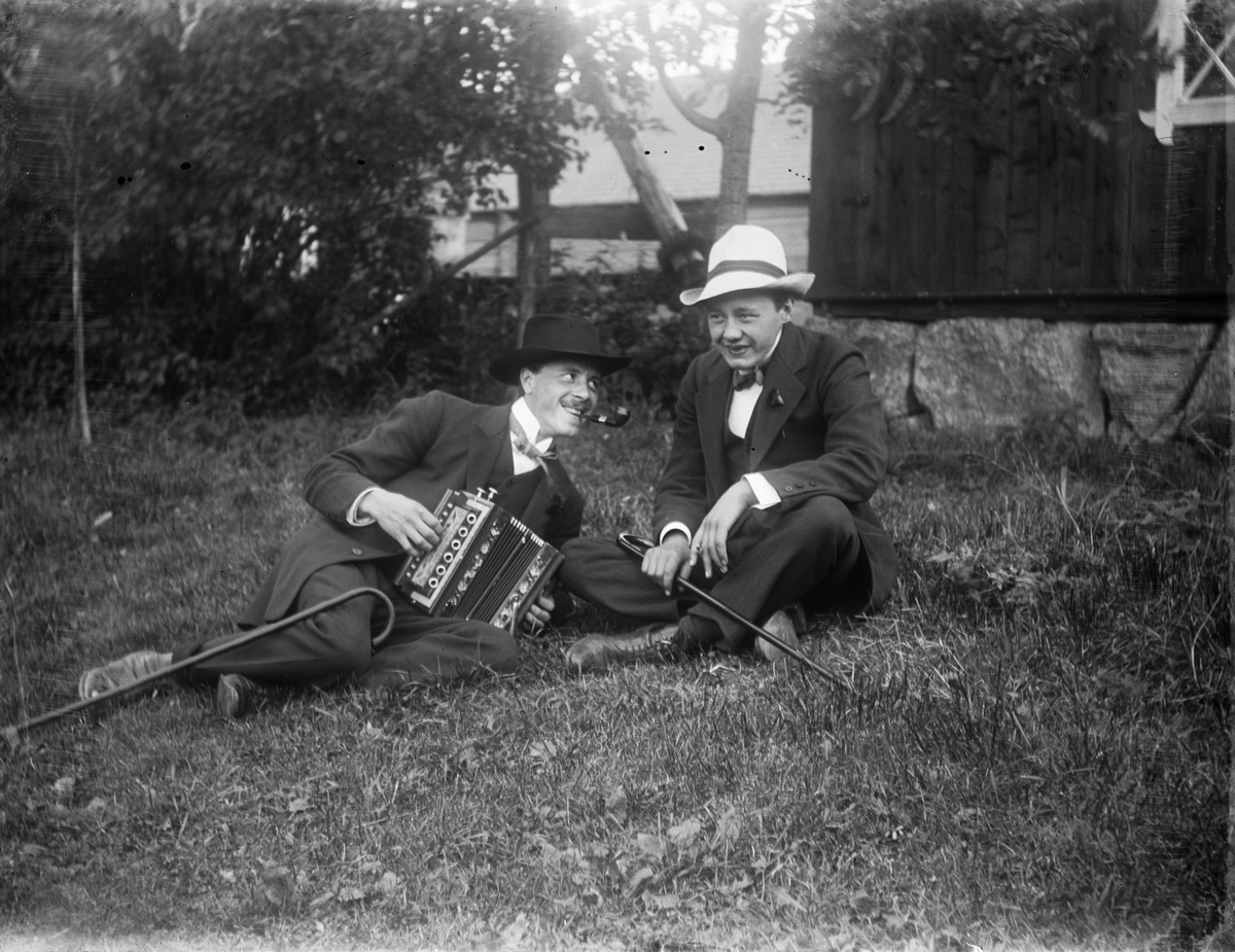 Dragspelande man och ytterligare en person sitter på gräset i trädgården, Uppland