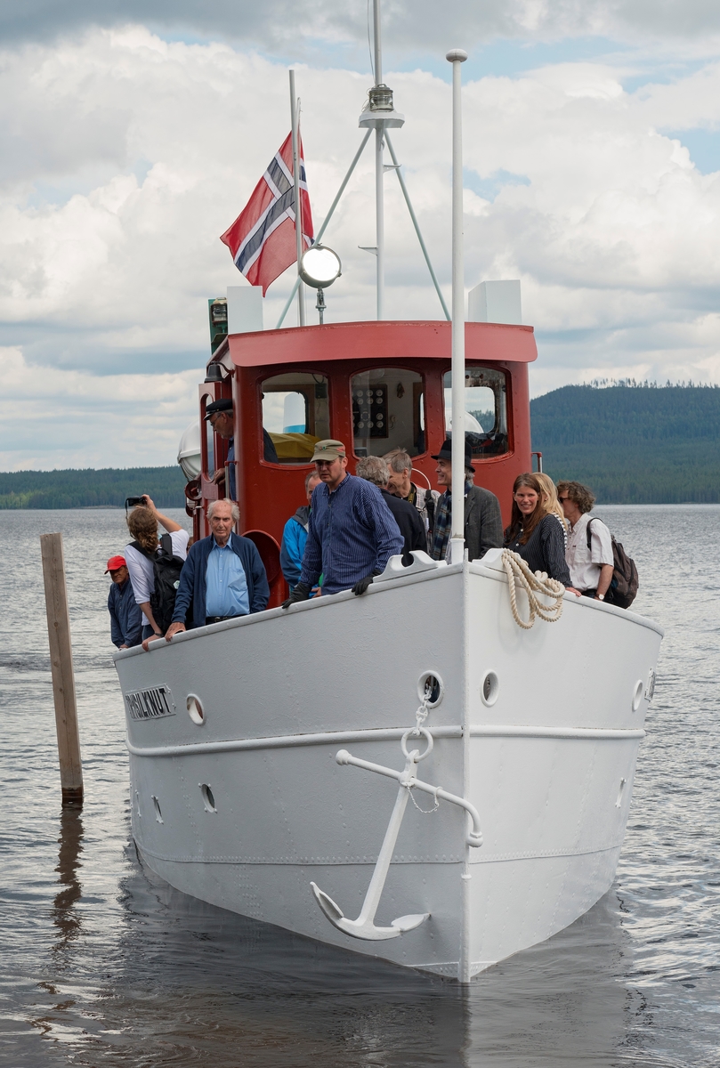Slepebåten «Trysilknut» fotografert på den 47 kvadratkilometer store Osensjøen i grensetraktene mellom Åmot og Trysil i Hedmark, der Trysilknut gikk som tømmersleper fra 1914 til 1984.  Etter den tid har den 55 fot lange og 12 fot brede båten vært brukt som museumsbåt.  Den ble restaurert i perioden 2011-2014, og dette fotografiet er fra den første turen båten gjorde med passasjerer etter at restaureringa var avsluttet med maling av eksteriøret.  Skroget er kvitmalt og overbygningen (styrhus og bysse) er rødbrun.  Disse fargene har Trysilknut hatt siden båten ble ombygd fra damp- til dieseldrift i 1957-58, bortsett fra at vulsten som markerer hvor rekka møter den nedre delen av skroget lenge var grønnmalt.  Fotografiet er tatt mot båtens baugparti idet den legger til ved brygga på Sørlistøa fløtermuseum etter en rundtur på sjøen som inngikk i markeringa av Trysilknuts 100-årsjubileum 11. juni 2014.  På fordekket ser vi blant annet pensjonert museumsdirektør Tore Fossum (som ledet Norsk Skogbruksmuseum da Sørlistøa og Trysilknut ble innlemmet i museet), maskinist og omviser Allan Tutvedt, avdelingsdirektør Stig Hoseth (bak flaggstanga) og fagkonsulent Ingvild Herberg, de to siste fra Norsk Skogmuseum. Mer om Trysilknut og dens historie under fanen «Andre opplysninger».
