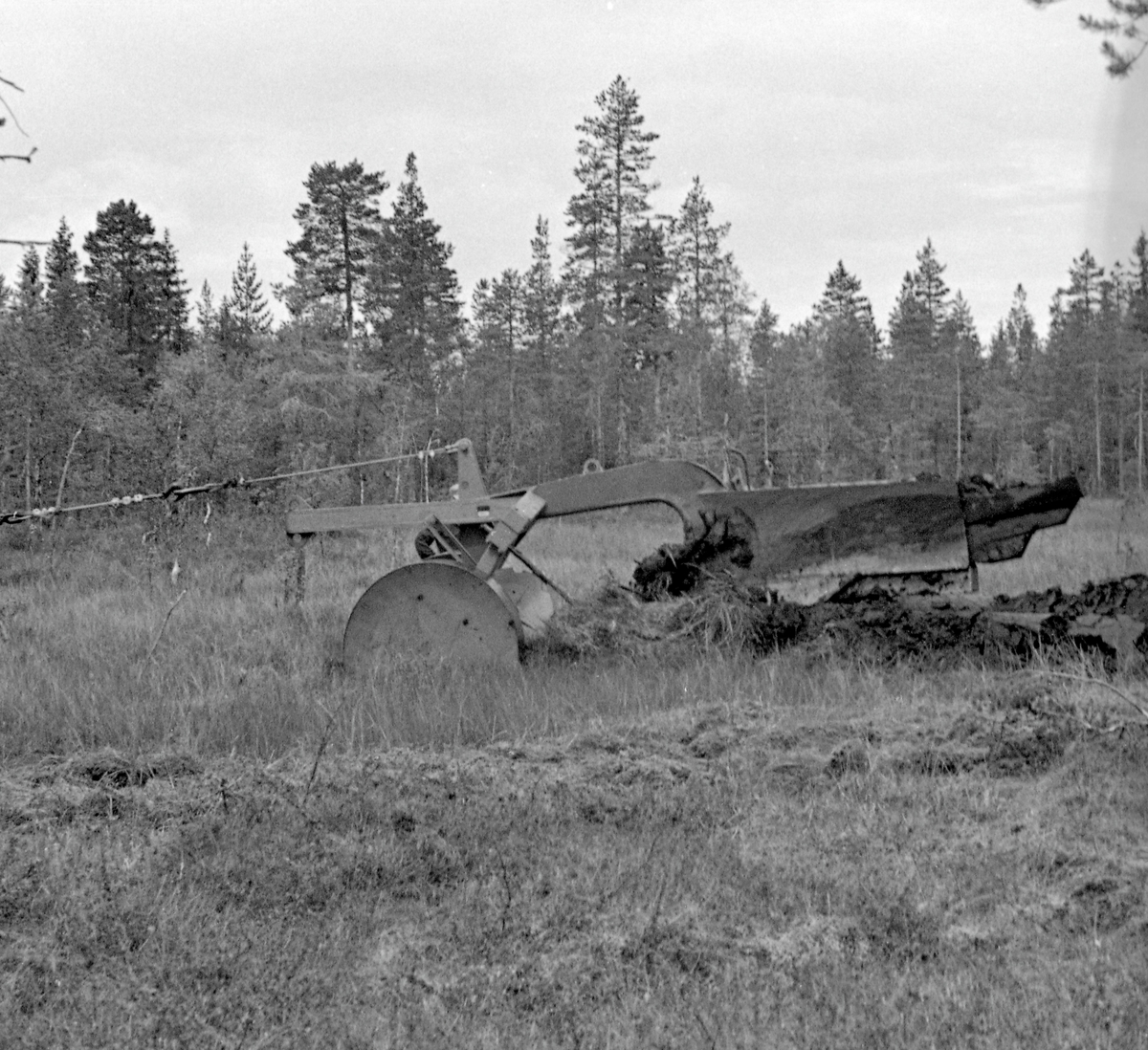 Grøfteplog, en såkalt «Lokomo-plog», fotografert under forsøksdrift på Grue Finnskog sommeren 1957.  Her ble både Langmoens skoger og familien Bredesen-Opsets skoger stilt til disposisjon for slike forsøksdrifter.  Grøfteplogen veide 3,8 tonn og ble trukket ved hjelp av en beltetraktor med bakmontert vinsj.  I front hadde plogen et par brede stålhjul, som skulle hindre at den grov seg ned i myra.  Derfra gikk det en kraftig, nedoverbøyd stålås med vridde veltefjøler, som veltet myrtorva opp og til side etter at den først var snittet av en sirkelformet kniv i forkant.  Lokomoplogen var robust, og kunne i mange tilfeller flytte stein, stubber og småtrær vekk fra grøftetraseen.  I noen tilfeller ble det imidlertid for mye.  Da ble festepunktet fra vaieren på vinsjen flyttet til den øvre delen av en vektarm, som fikk plogen til å heve seg fra myra.  Deretter kunne plogen senkes igjen ved å flytte vaierfestet til den nedre delen av vektarmen igjen, slik vi ser det er gjort på dette fotografiet.  Da dette bildet ble tatt sto to menn i samtale bak plogen.  Mer informasjon om Lokomo-plogen finnes under fanen «Opplysninger».