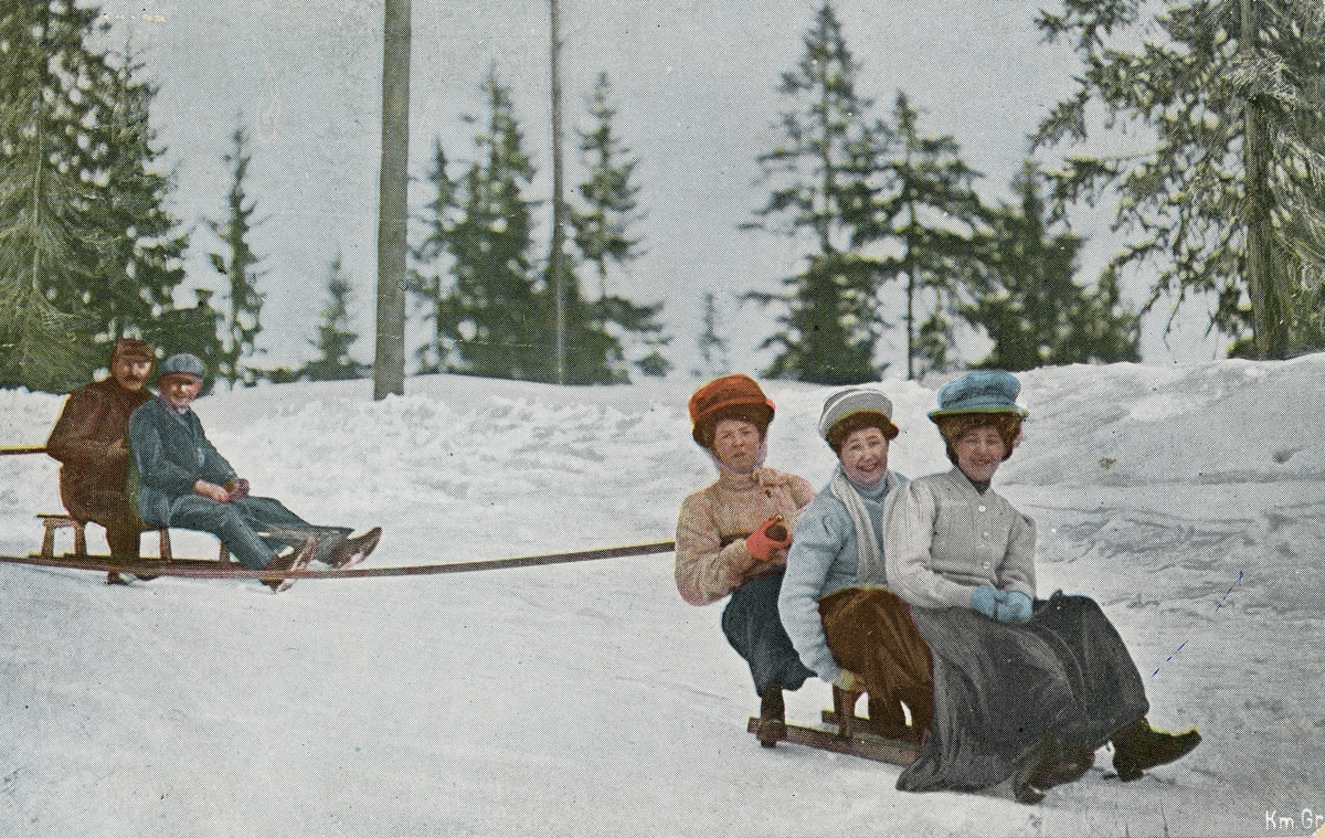 Postkort. Det håndkolorerte fotografiet på kortets fremside viser menn og kvinner på aketur i vinterlandskap.