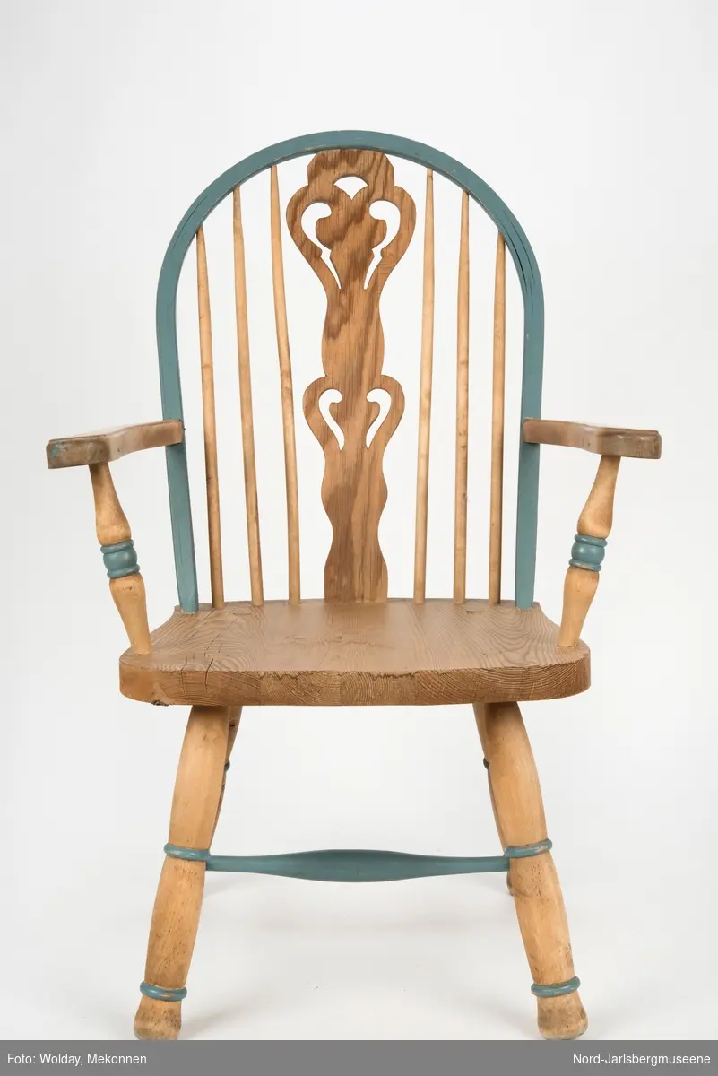 "Budal"inspirert stol, med ryggstø og armelner. Ryggstø er formet av en blåmalt buet list som holdes oppe av seks vertikale trespiler. Den midter, og syvende, spilen er en bred, utskjært list med gjennombrutt mønster, stiliserte hjertemønster. Armlenene er buet utover, og ben og detaljer har dreid dekor.