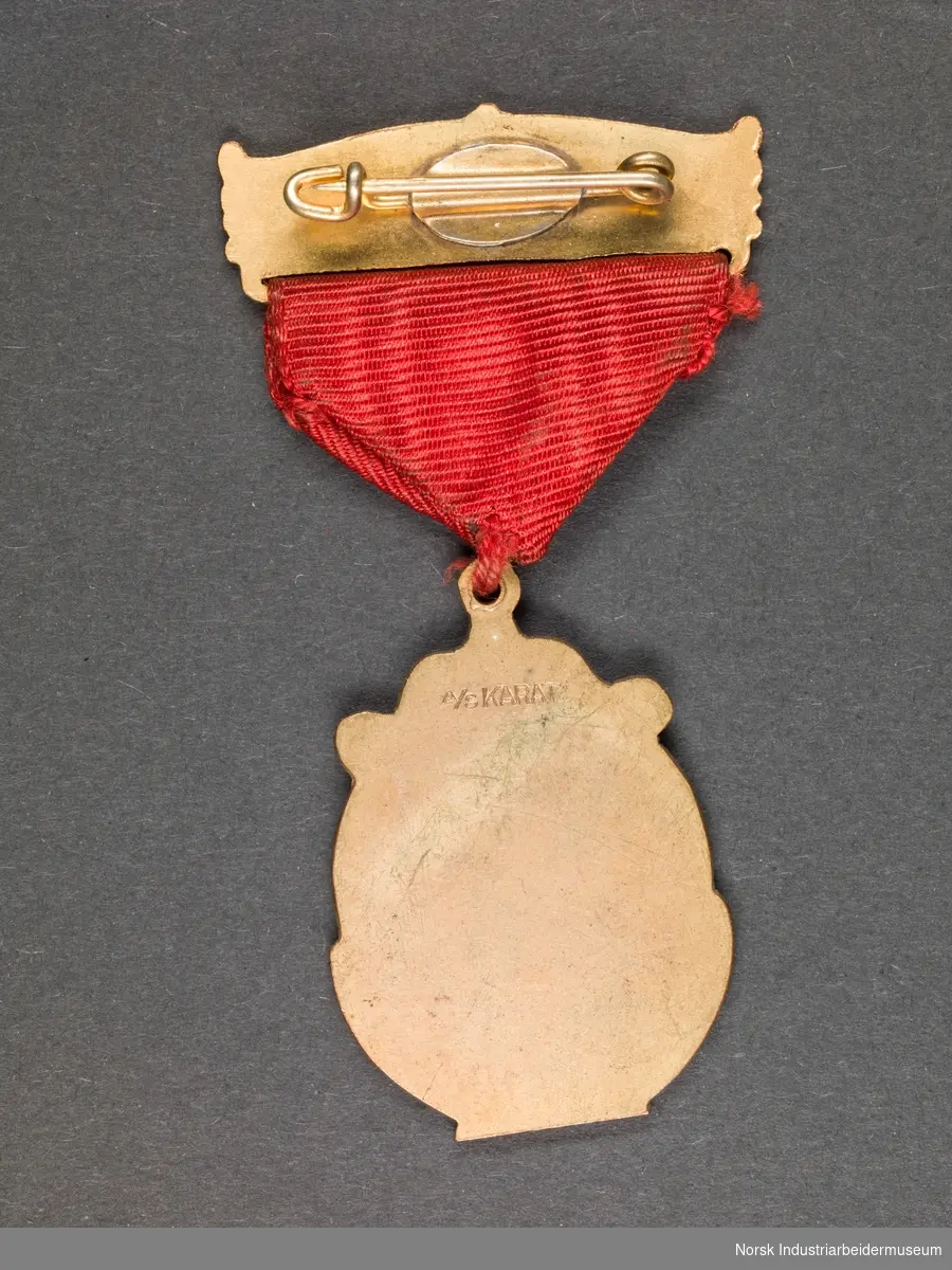 Gullfarget medalje festet til rødt tekstilbånd og gullfarget nålfeste. Motiv på medaljen er en kirke. Nålfestet er dekorert med en note.
