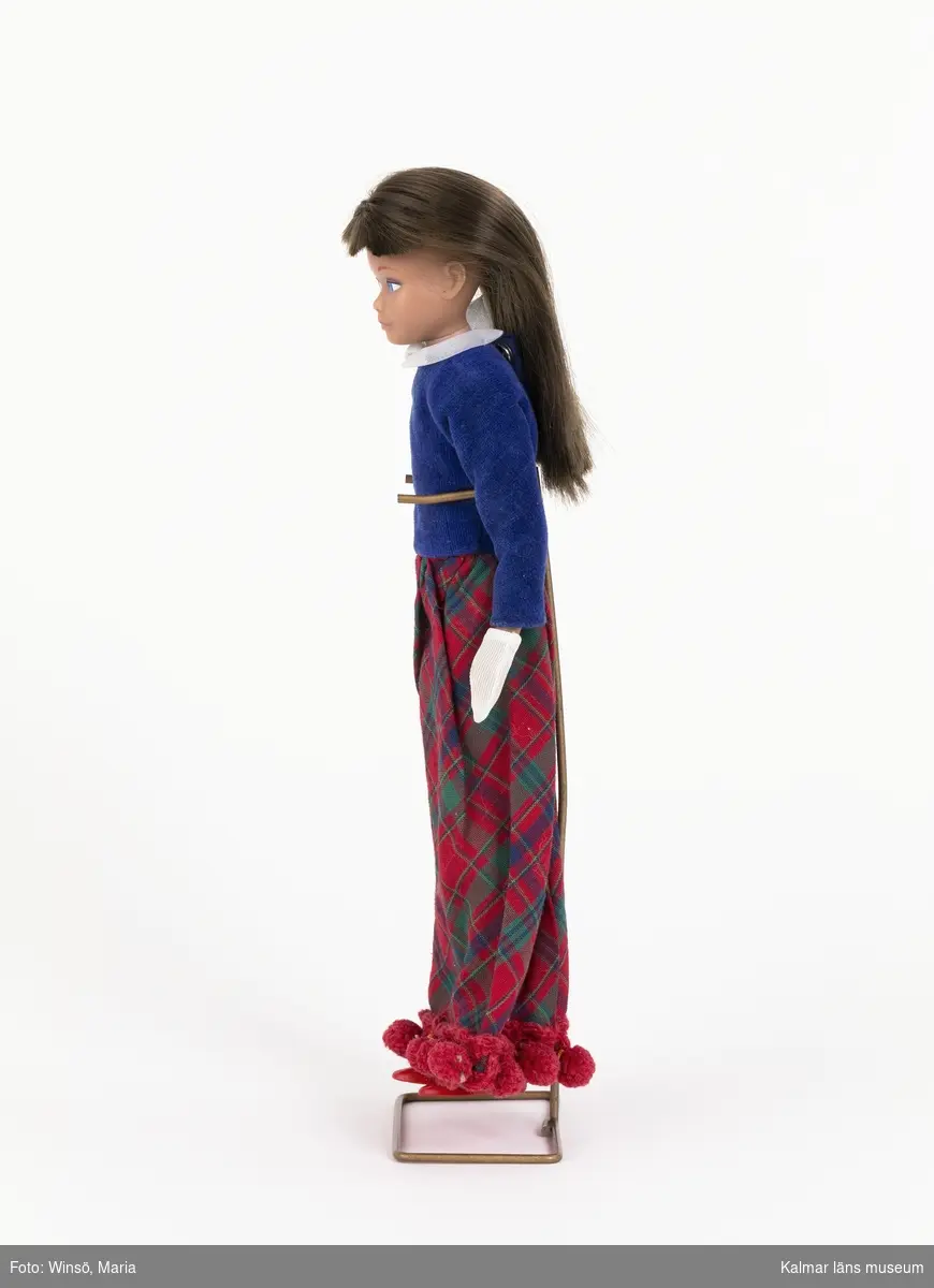KLM 45643:1 Docka, Skipper med klänning, handskar, strumpor, skor, cape, skivspelare, ställ och  originalkartong. Skipperdocka som förvaras i en halv originalkartong där dockan ligger i en metallställning.  Ställningen är till för att dockan ska kunna stå. Dockan har brunt långt hår. Den är klädd i en klänning med blått, långärmat blusliv med vit krage, kjol med rutmönster i blått och grön samt röd bollfrans i fållen, vita handskar, vita strumpor, röda ballerinaskor av plast med texten ”JAPAN” på sulan, samt skivspelare av blå och vit plast, skadad genom att någon tuggat på den. Klänningen märkt med Mattels etikett. Skivspelare i plast. I ena hörnet har familjens katt tuggat på plasten. Setet finns i häftet ”Barbie och hennes vänner – med många nya kläder” från Brio och heter ”914 Skivparty”. Dockan har dessutom en hemtillverkad röd cape som är tillklippt ur ett rött filttyg och försedd med tryckknapp. I capen har ett stycke klippts bort. Den kvarvarande delen av kartongen har tryckta bilder av Skipper med ljust, svart eller brunt hår samt olika kläder.  På ovansidan står texten: Barbies Little sister Skipper by Mattel. Skipper has her own ”junior-edition” styles…They match Barbies wonderful warderobe. På en av kortsidorna står “ Stock No 0950 (Redhead).