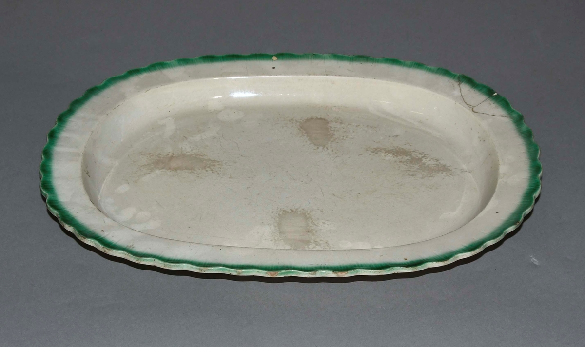 Ovalt stekefat av hvit keramikk. Kanten er bølgete og har en grønn dekorborde.