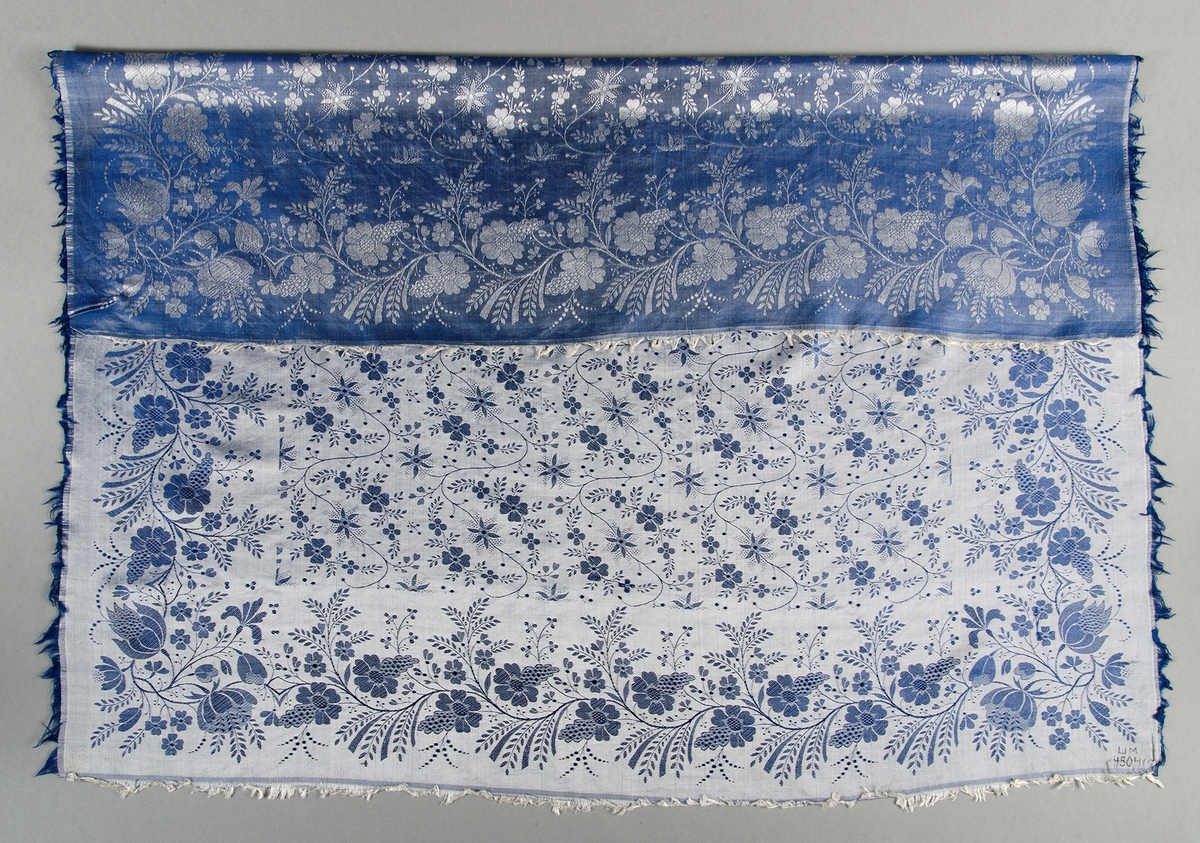 Kläde av siden med vävt blommönster i blått och vitt. Korta blå och vita fransar av varp och inslag. Stockholms hallstämpel 1842.
