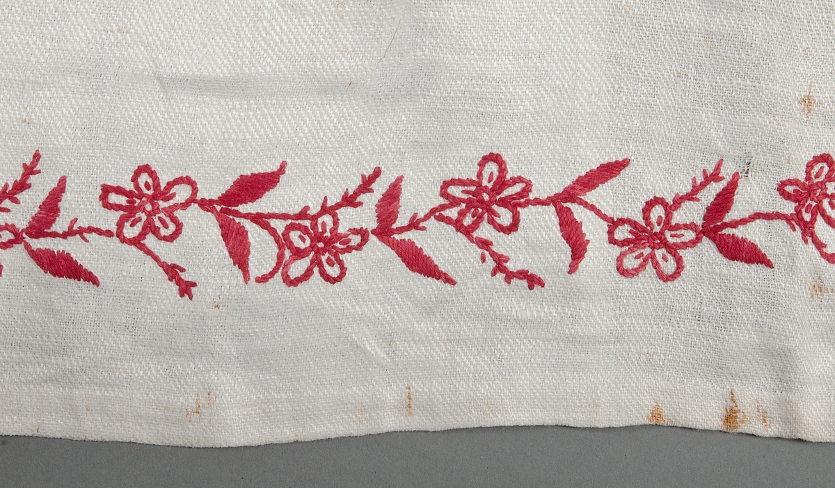 Av vitt kypertvävt linne. Blomranka broderad med plattsöm och stjälksöm i rött bomullsgarn. Förklädesband av vitt bomullsband.