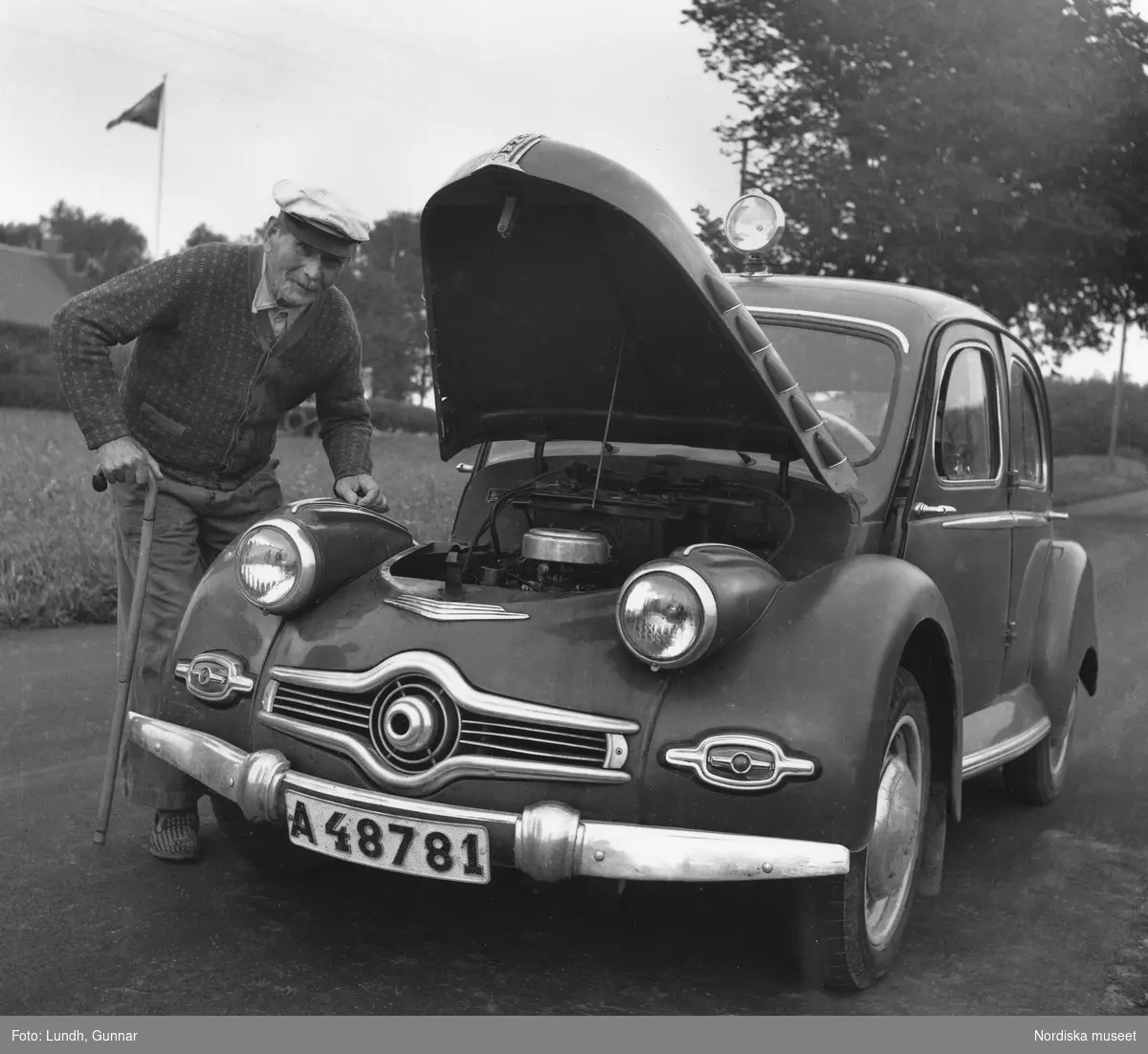 Fotografen Gunnar Lundhs bil, en Dyna Panhard med uppfälld motorhuv. Äldre man i skärmmössa och med käpp står intill.