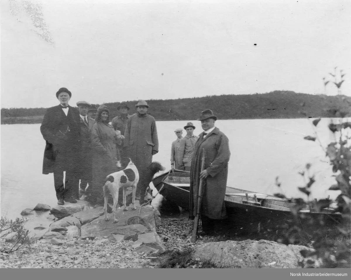 En gruppe mennesker avbildet i vannkant, med båter og hunder.