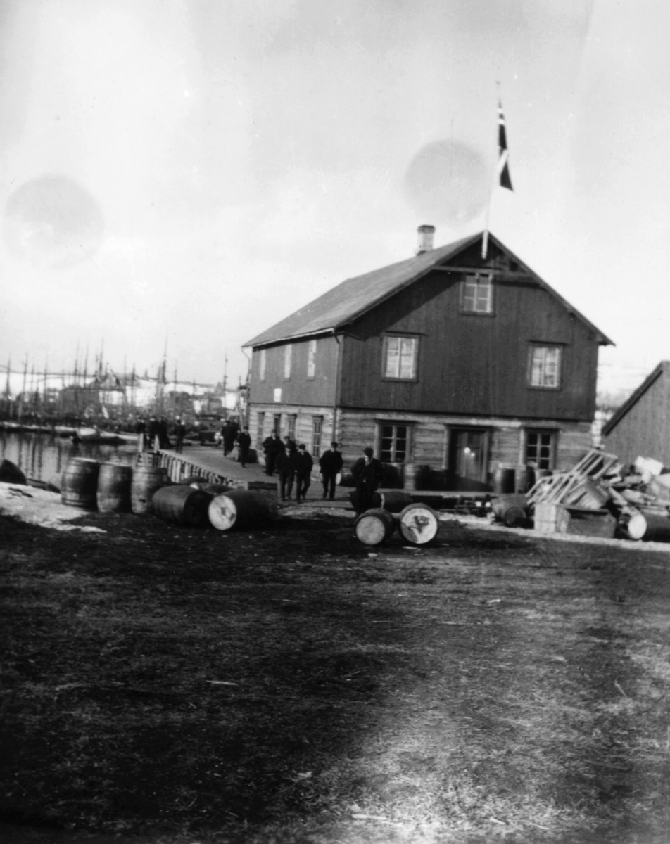 Menn på kaia ved et pakkhus i Båtsfjord i 1910. Flere tønner ligger utenfor byggningen. Havna er full av fiskebåter. Det er vår og det norske flagget er heist opp på toppen av byggningen.