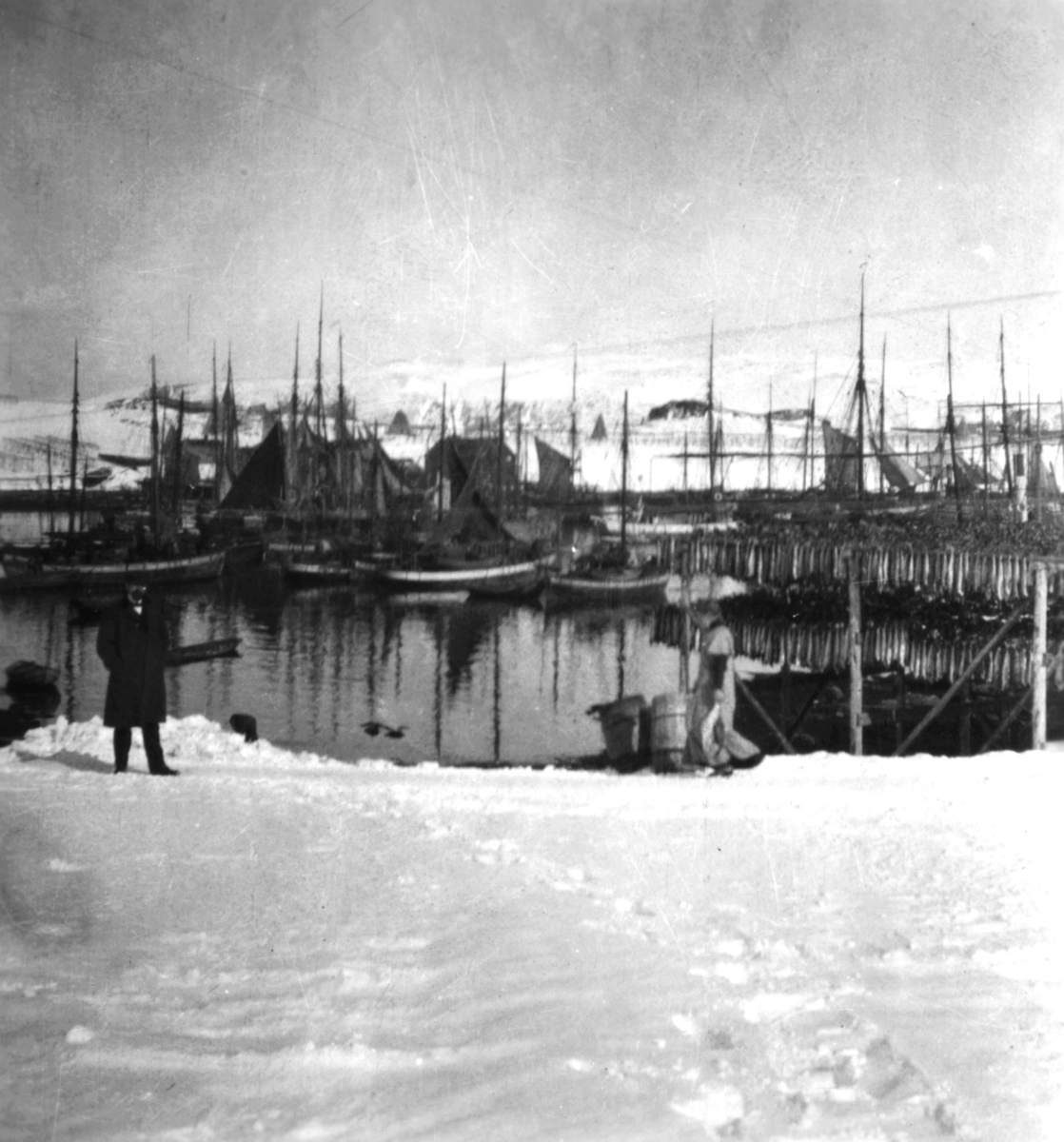 Postekpeditør Rasmus Kyvik står til venstre ved Båtsfjord havn. En mann går på stien i snøen og bærer fisk til eller fra hjellene. Flere fiskebåter ligger ved havn.