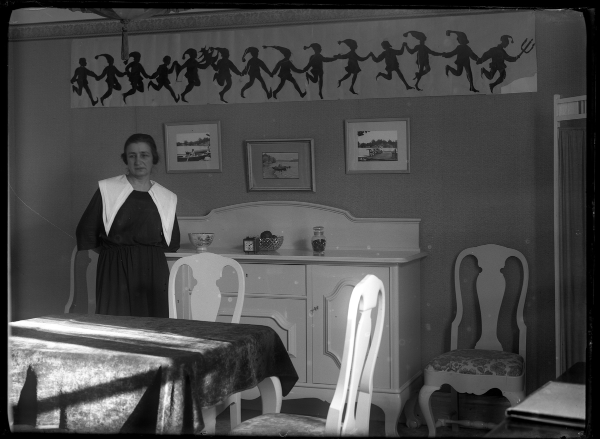 Anna Lenander står lutad mot en vit skänk i en matsal. På väggen hänger en pappersbonad med tomtenissar i dans.