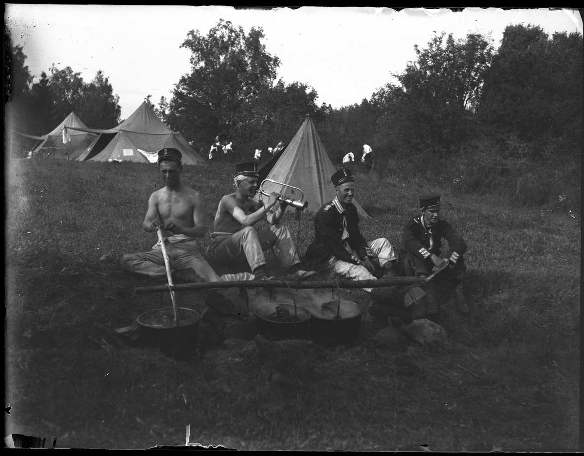 Gruppbild tagen under fotografen Harald Olssons excercistid. Vid ett läger, framför tre tält, sitter fyra män och förbereder mat. De är alla klädda i olika uniformsdelar och den ena spelar på ett blåsinstrument.