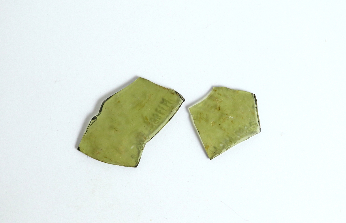 Två fragment av en cylindrisk och något bukig bägare av ett gröntonat glas.