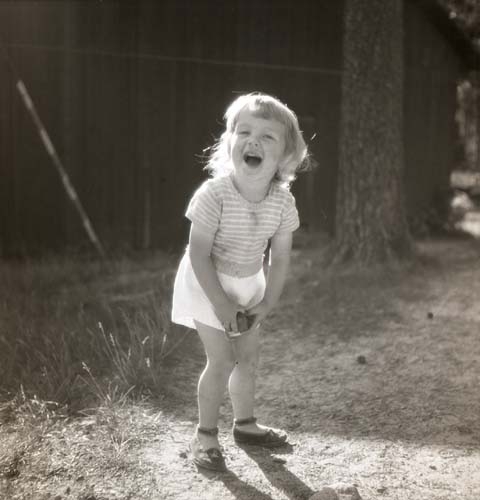 Ett barn iförd shorts, kortärmad tröja och sandaler tittar skrattade mot fotografen. I händerna håller barnet en sten, 1951.