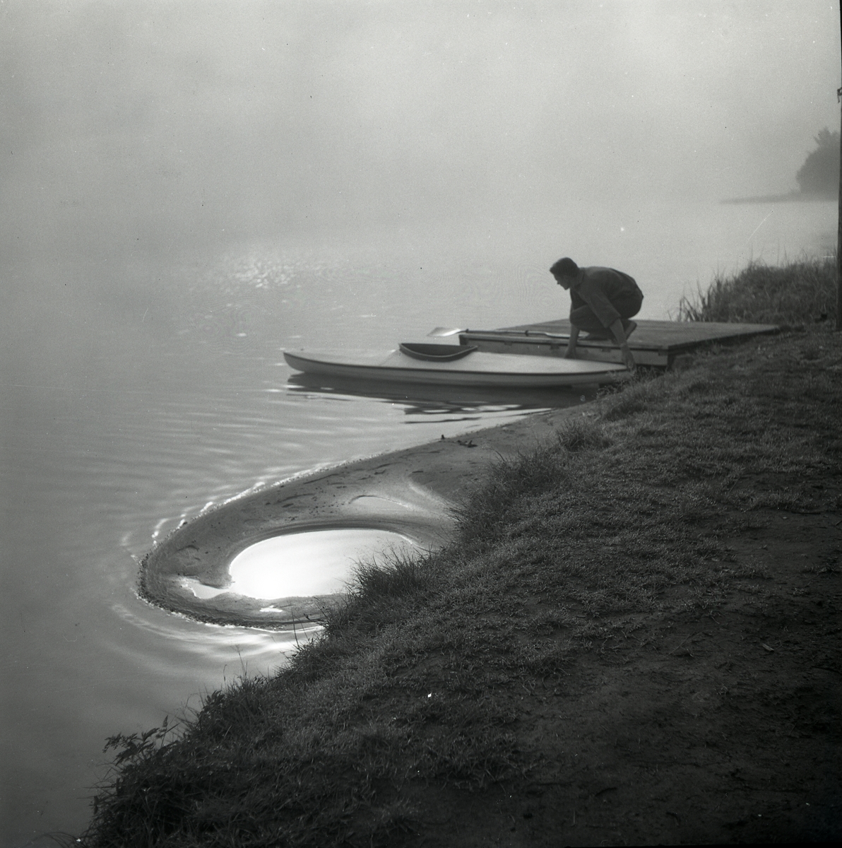Hilding Mickelsson sitter på huk på en brygga vid en kanot. Det är en disig  morgon vid Skidtjärn i augusti 1949. Solen glittrar i vattnet.