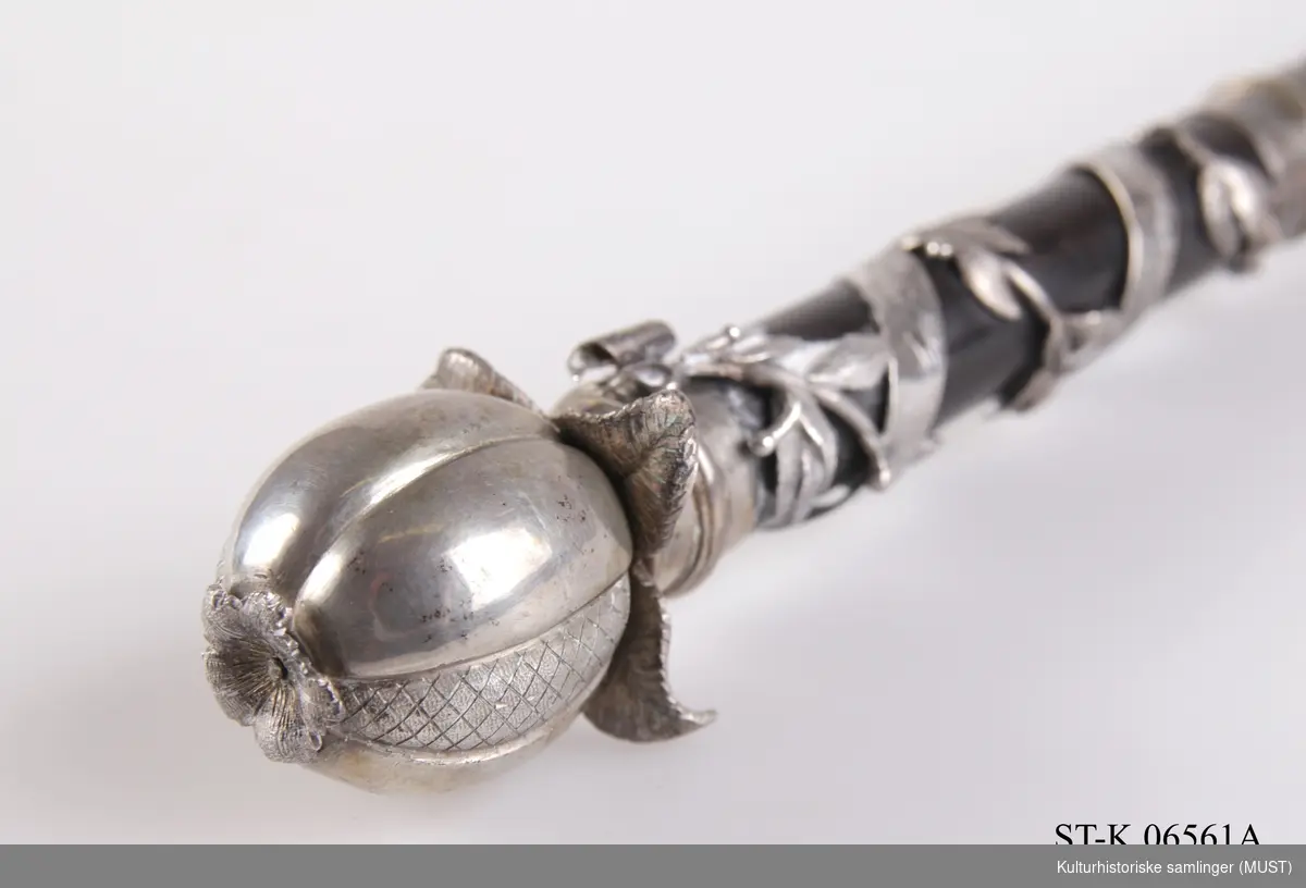 Dirigentstav dekorert med et granateple i sølv på toppen. Håndtaket er omviklet med en lauvbærranke og et innskriftsbånd i sølv.