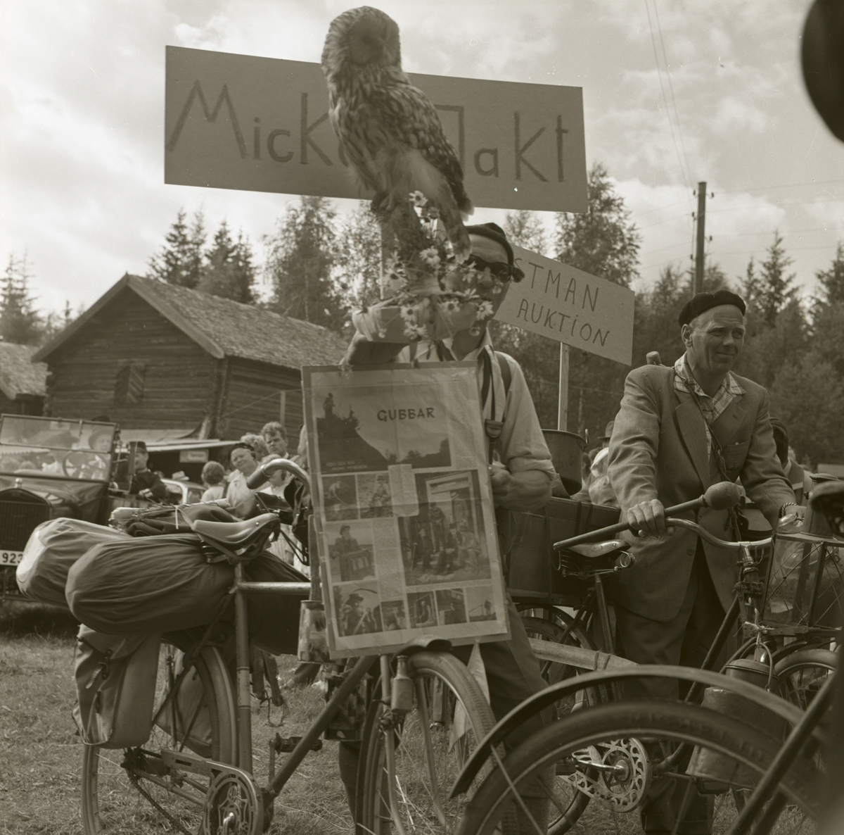 Karneval under en hembygdsfest där två män leder varsin cykel med packning på pakethållaren samt har varsin banderoll med text och bild på. Publiken tittar nyfiket på ekipaget, 22 juni 1962 i Rengsjö.