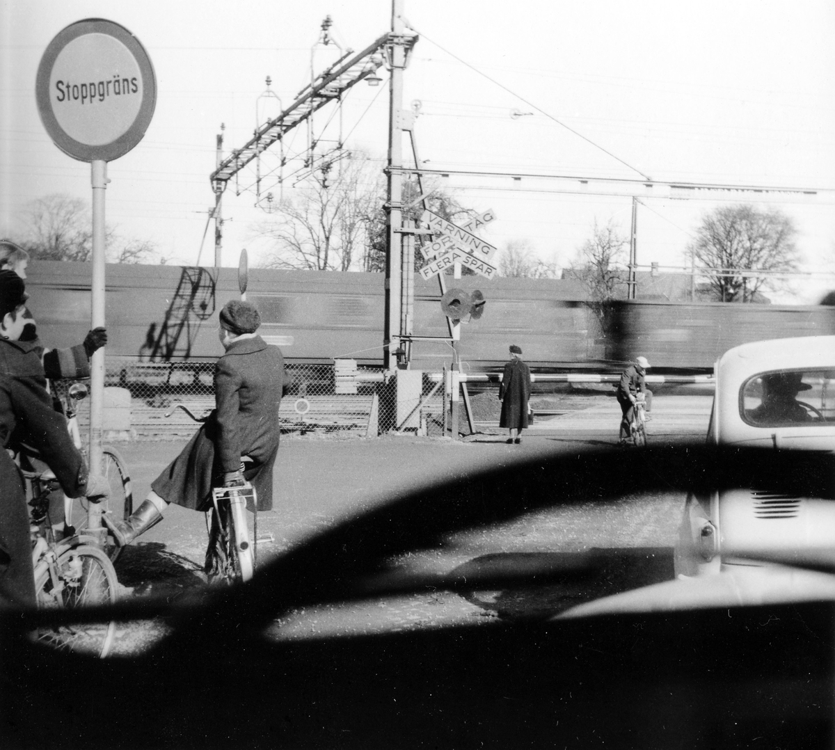 Flera personer, gående, cyklister och bilister, står vid en järnvägsöverfart och väntar på att ett tåg ska passera. Bilden är tagen från framsätet av en bil.
Järnvägsöverfarten byggdes bort i slutet av 1980-talet.