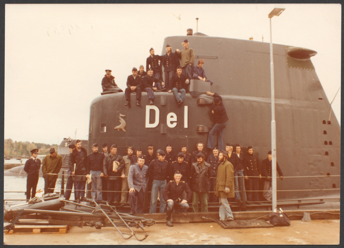 Grupporträtt som visar krigsbesättning för ubåten Delfinen under en repetitionsövning 1972. En del av manskapet är uppställt framför ubåtstornet som dominerar bilden. En annan del sitter uppe på tornet. Mannen som håller på att klättra upp på sidan är givaren Lars Söderlind.