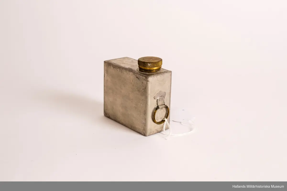 En oljeflaska med skruvlock, tillverkad i tenn. Med ring på ena kortsidan. Locket är märkt på ovansidan "PRIMUS".