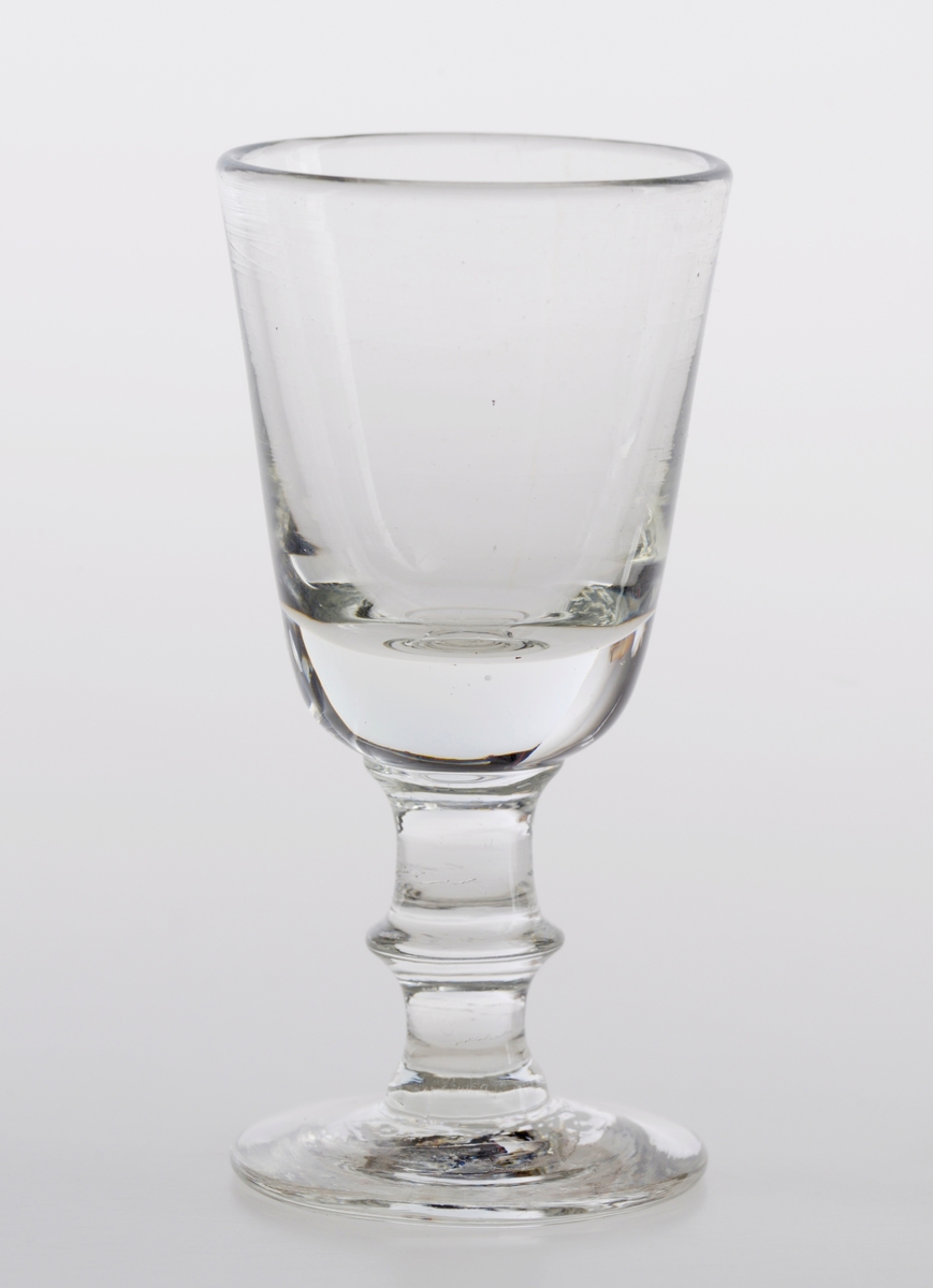 To små drammeglass laget av klart glass. Klokken er høy og relativt smal. Munningen er større enn foten. På stetten er det dekor i form av en ring av glass midt på foten.