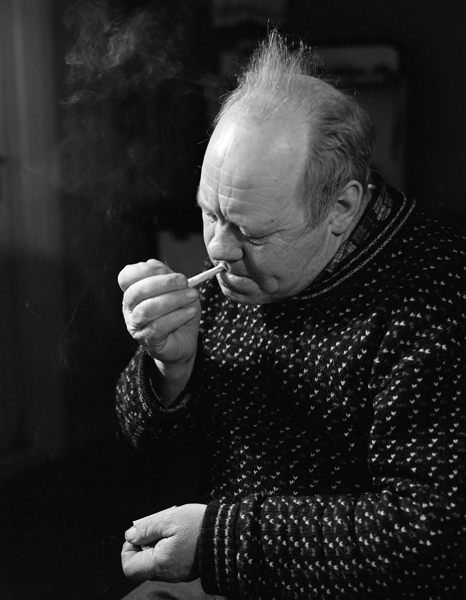 Oskar Martinussen, Fåberg, tenner en sigarett.  Fotografiet ble tatt på Martinussens kjøkken, der medarbeidere fra Norsk Skogbruksmuseum dokumenterte hvordan han bandt en fangstpose til grunnhåv.