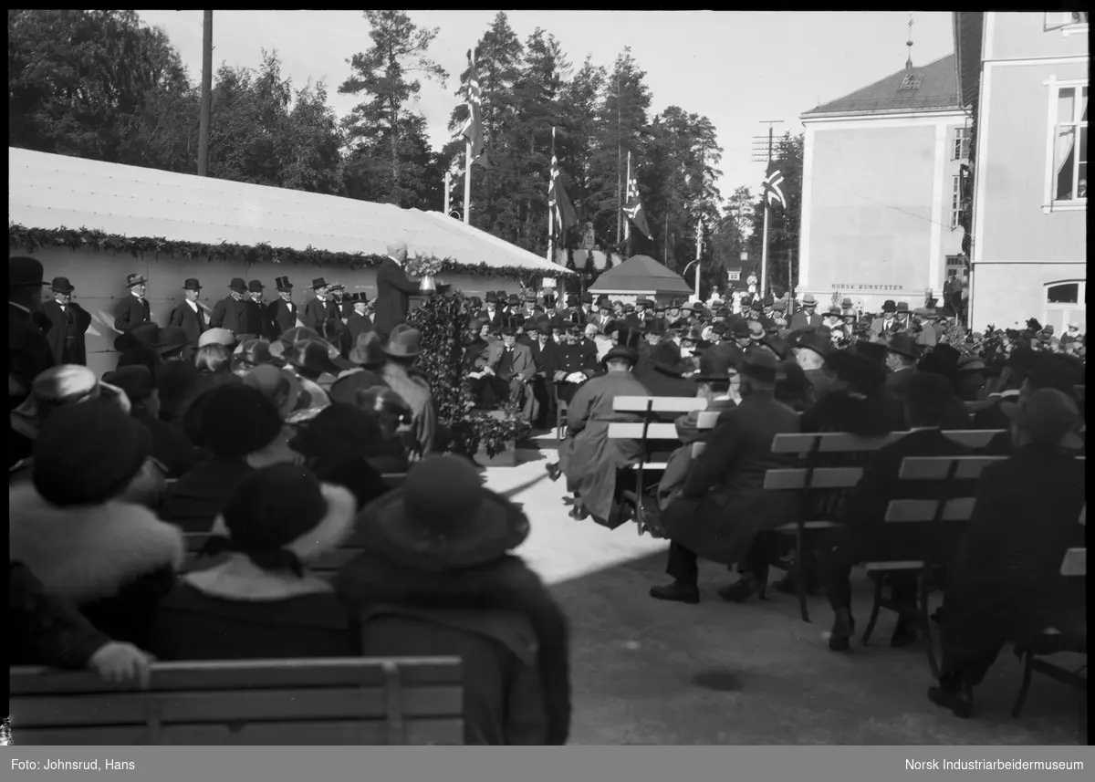 Åpning av Fylkesutstillingen 1922 med besøk av Kong Haakon VII. Person taler foran folkemengde. HM Kongen sitter som tilhører.