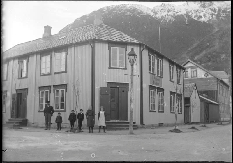Personer og barn utenfor forretningen P. Nygaard. På hjørnet C.M. Havigs gate og Petter Dass gate. Øyfjellet i bakgrunnen.