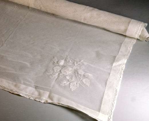Kläde av vit bomullstyll kantad med 1 cm bred knypplad spets runt fållen. I ena hörnet trätt blombroderi i vitt.