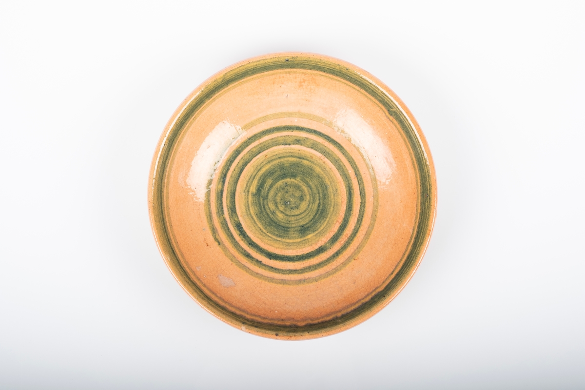 Glasert, rund keramikkskål. Skålen er dekorert med grønne sirker, som utgår fra midten, på rødorange bunn.