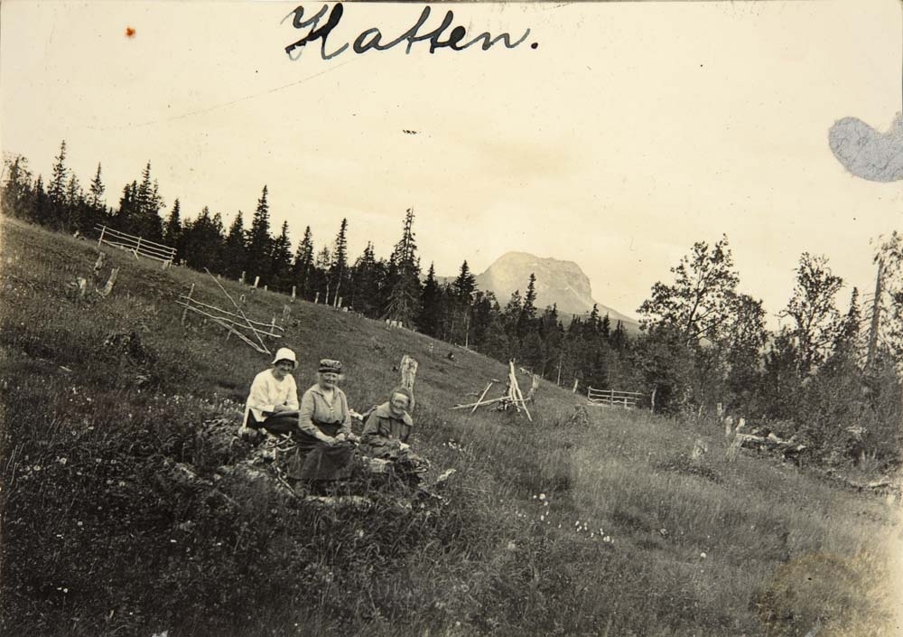 Tre kvinner sitter i en eng i åsen. Hatten skimtes i bakgrunnen.