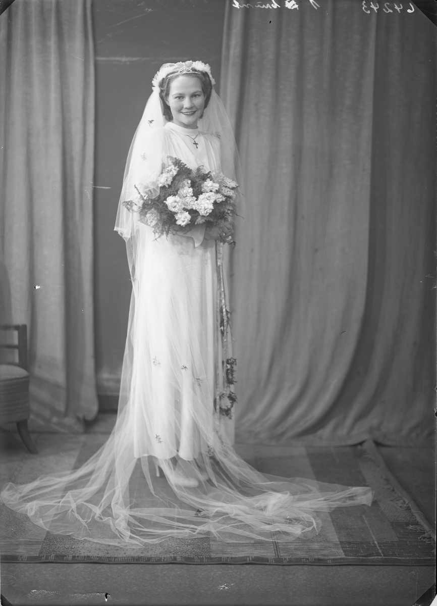 Gruppebilde. Brudebilde. Ung lyshåret kvinne i lang hvit brudekjole med hvit slør og en blomsterbukett i armene. Brud. Bestilt av Hr Carl Ivar Ørgland. Vikebygd