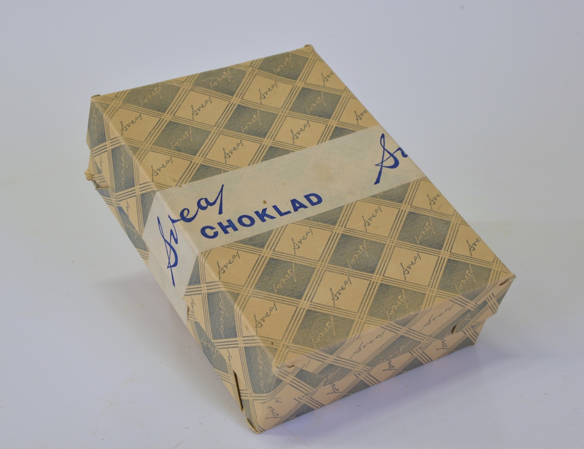 
Rektangulär ask med rutigt mönster i vitt och blått. I varje ruta texten "Svea". Vit klisterremsa tvärs över asken med texten "Svea choklad" i blått.
Från Arne Ekströms handelsmuseum.