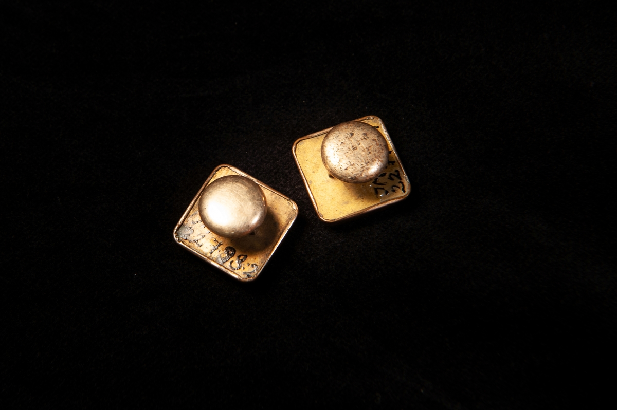 Fyrkantig stolpknapp av förgylld metall (gulddoublé) med präglad dekor i form av två tvärrandiga ovaler med en silver- och en guldfärgad blomma. Samhör: JM 22798:1-2.