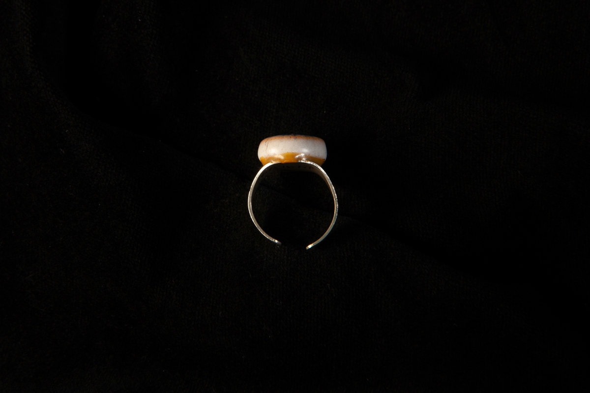 En ring av metall (silver?) med en sten av glaserat lergods i blått och vitt. Inte sluten ring. Ligger i en vit plastask med rosa bomull i.