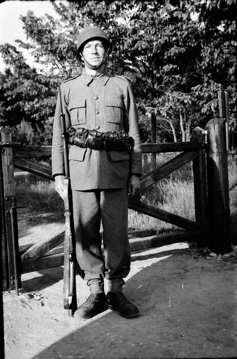 En militär, sannolikt en beredskapsman under andra världskriget, står på vakt med gevär och bälte (troligen magasinsfodral) vid en grind.