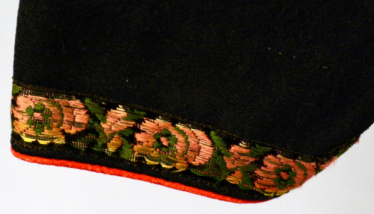 Kvinnetrøye av svart klede, kanta med kjøpeborder i mønster av raudt og grønt på svart botn. Høg krage, ope bryst. Fra protokoll.